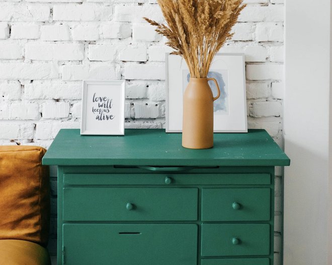 a bright green chest of drawers with a couple art prints and vase of dried flowers against a white brick wall